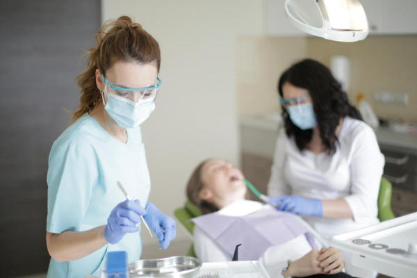 A dental practitioner wearing a face mask and protective glasses, attending to a patient in a dental chair with another staff member assisting in the background.