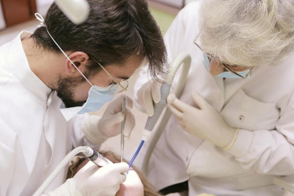 A dentist in a face mask and protective glasses attending to a patient in a dental chair with another dental practice staff member assisting them.